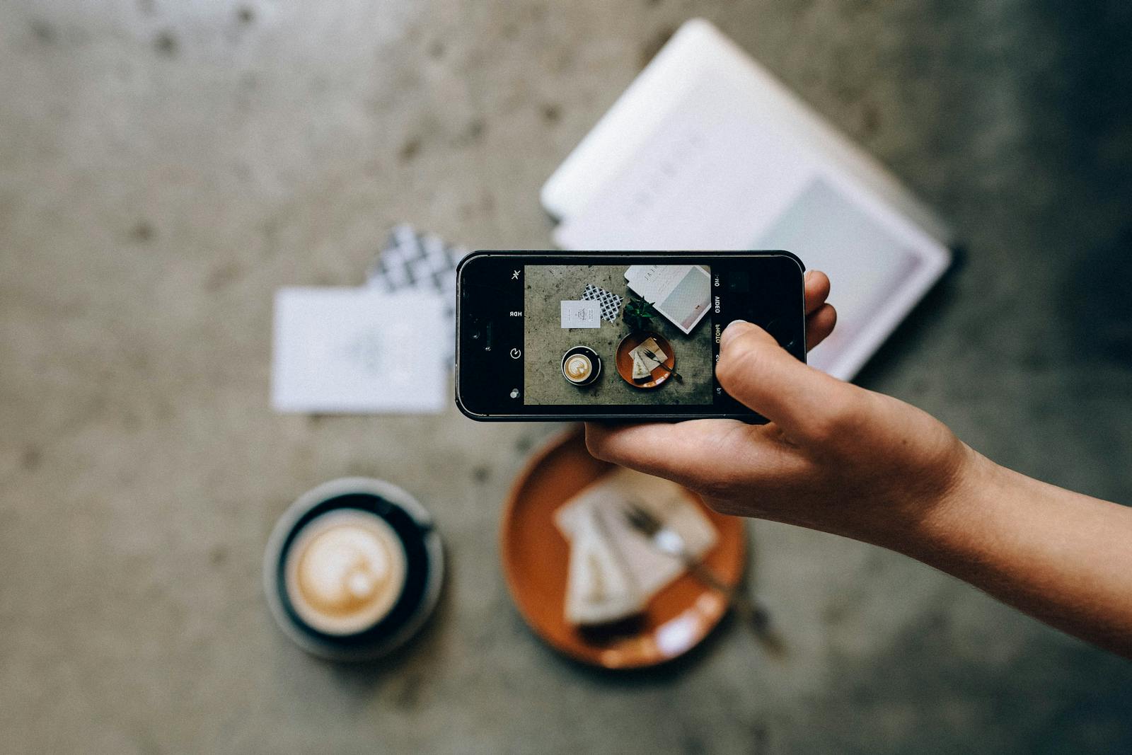 Hand hält Smartphone und fotografiert Kaffee und Kuchen von oben für Social Media.