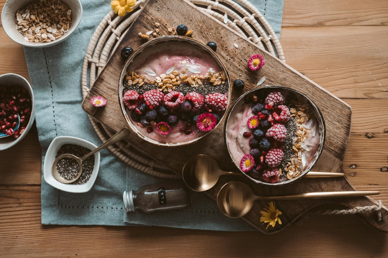 Flatlay von zwei Smoothie-Bowls mit Beeren, Granola und Kokosflocken auf rustikalem Holzbrett.
