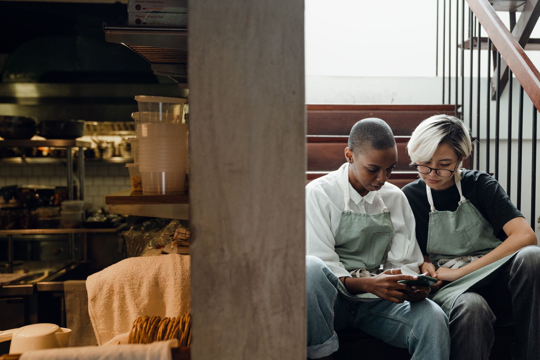Zwei Mitarbeiterinnen in Schürzen schauen gemeinsam auf ein Smartphone neben der Restaurantküche.
