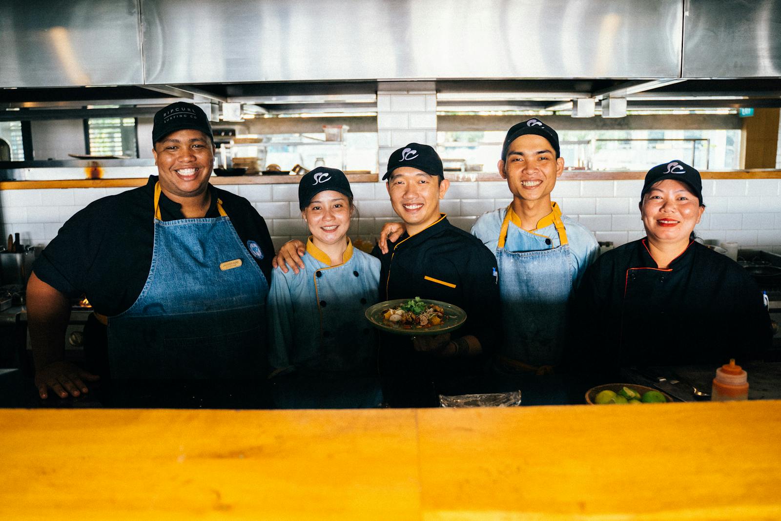 Lächelndes Restaurant-Team aus Koch und Servicekräften in offener Küche.